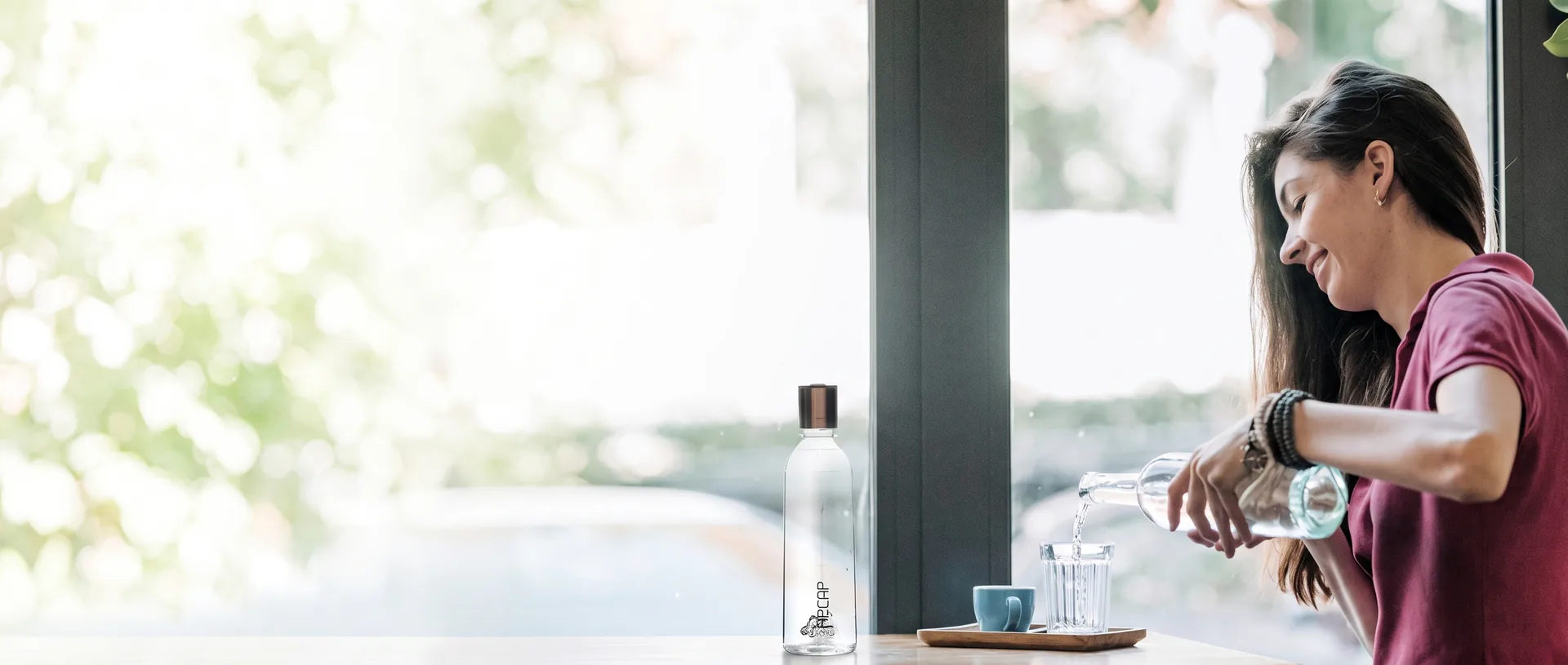 Woman pouring hydrogen water from an H2CAP PLUS bottle into a glass by a bright cafe window